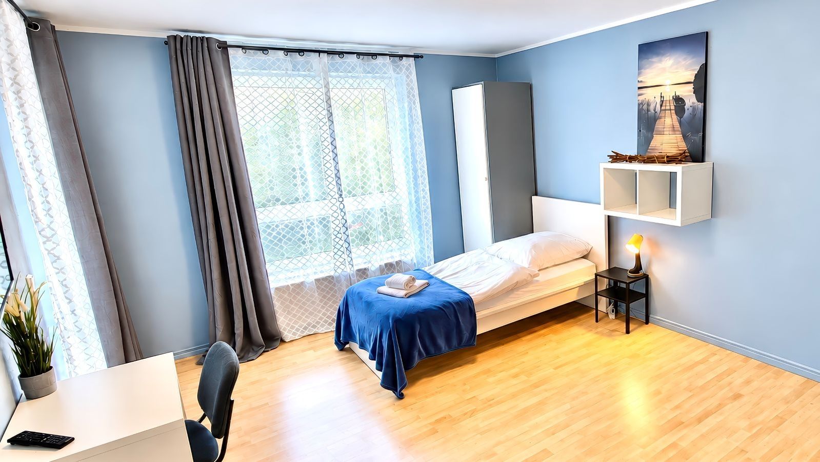 Schlicht eingerichtetes Schlafzimmer mit Einzelbett, blauem Bettüberwurf, Schreibtisch, Stuhl, Gardinen vor großem Fenster und Wandbild über Nachttisch mit Lampe