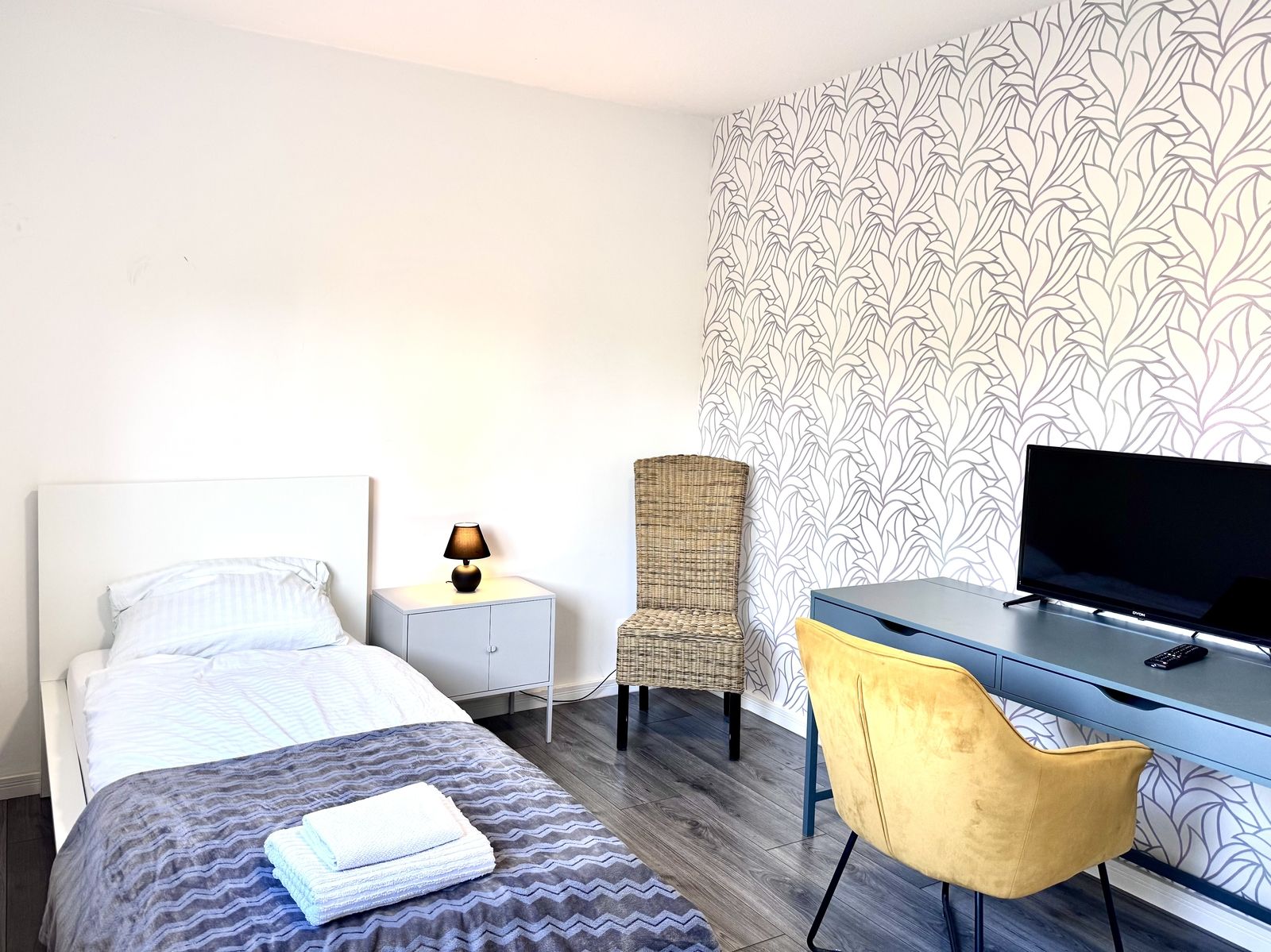 Gemütliches Einzelzimmer mit Einzelbett, Nachttisch, Schreibtisch mit gelbem Samtstuhl und Fernseher vor gemusterter Akzentwand