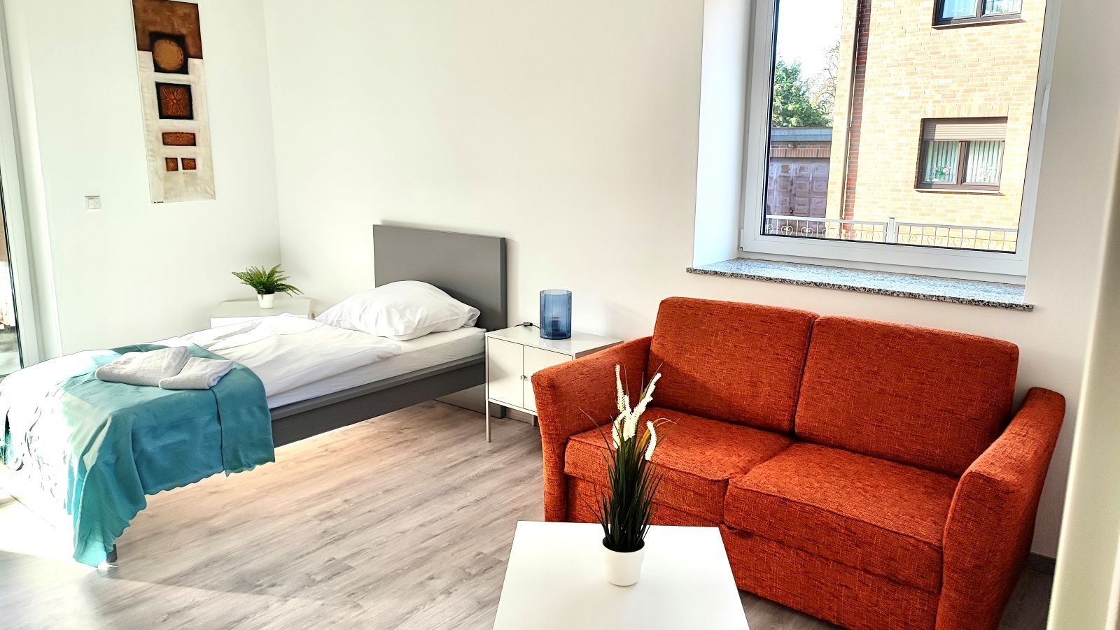 Helles Zimmer mit Einzelbett, rotem Sofa, weißem Beistelltisch und Fenster mit Blick auf Nachbargebäude