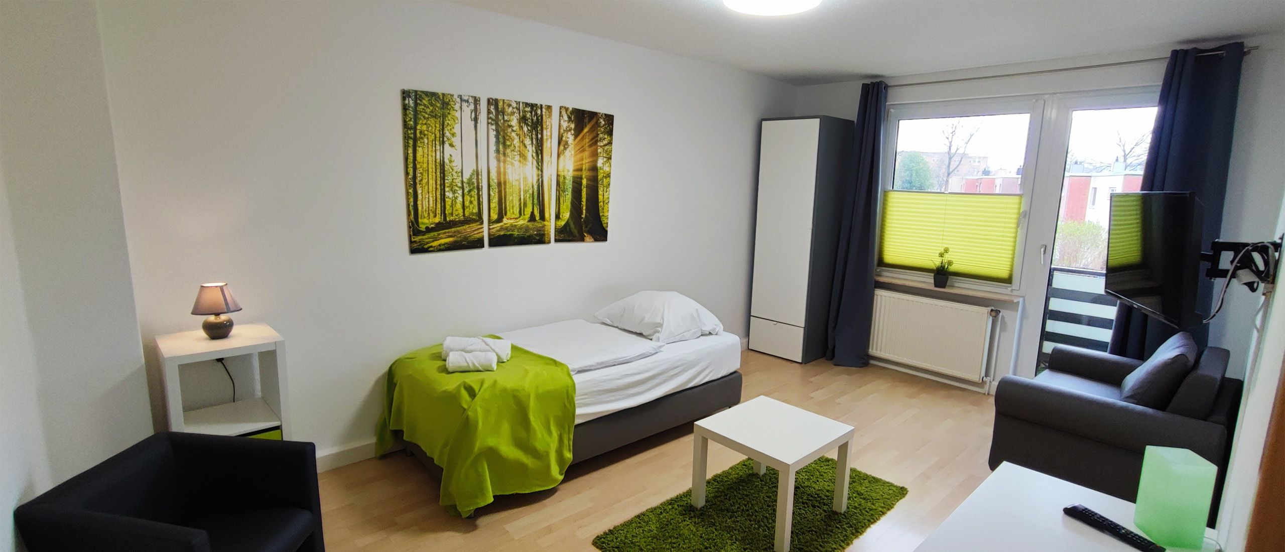Kleines Zimmer mit Einzelbett, grünem Überwurf, grünem Teppich, Sofa, Sessel, weißem Tisch, Nachttisch mit Lampe und dreiteiliger Waldwanddekoration