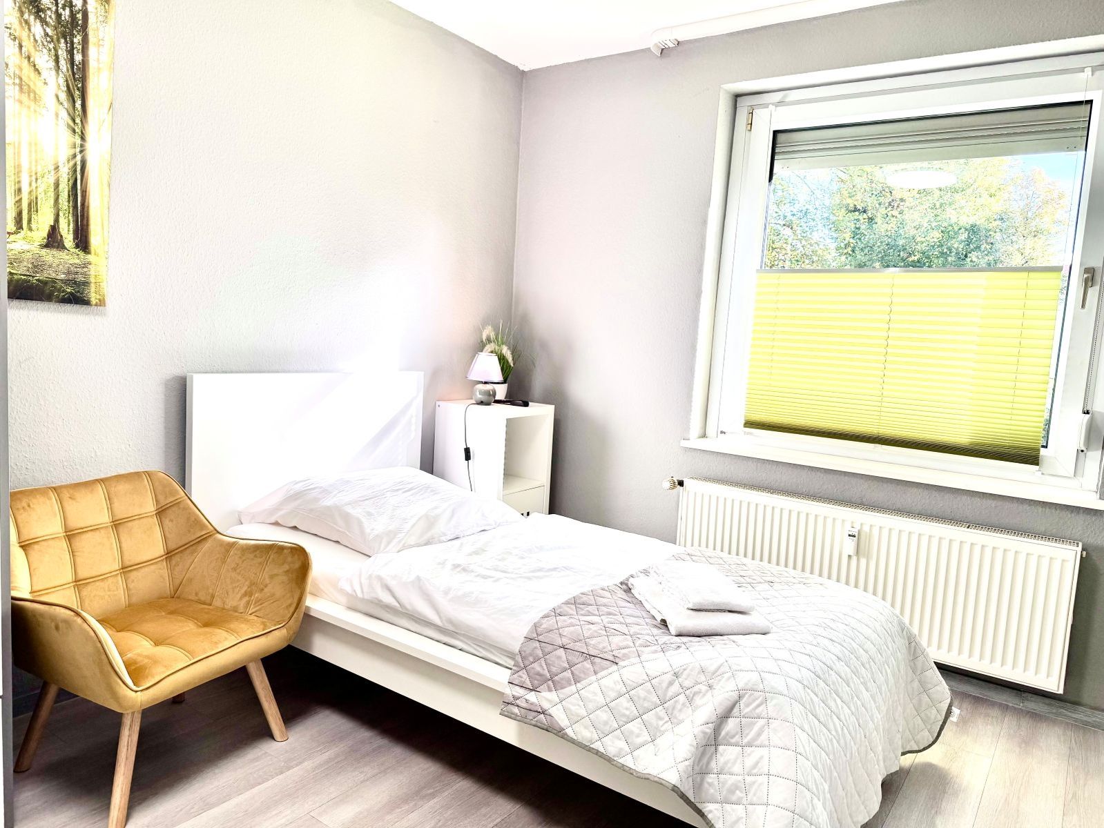Schlicht eingerichtetes Schlafzimmer mit Einzelbett, gelbem Sessel, Nachttisch mit Lampe und Fenster mit gelbem Plissee