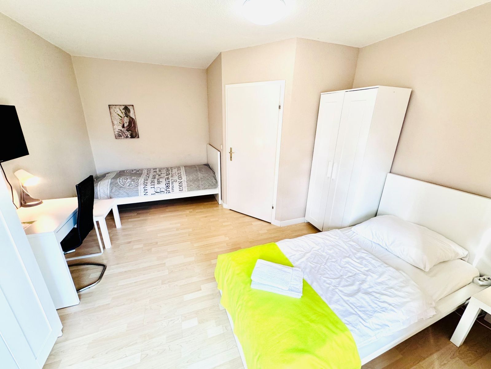 Schlicht eingerichtetes Schlafzimmer mit zwei Betten, einem Schreibtisch mit Stuhl, einem Kleiderschrank und einem Bild an der Wand