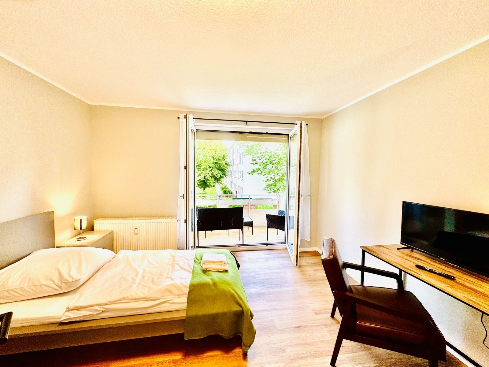 Helles Schlafzimmer mit Einzelbett, grünem Überwurf, Schreibtisch mit Stuhl und offenem Balkon mit Blick ins Grüne