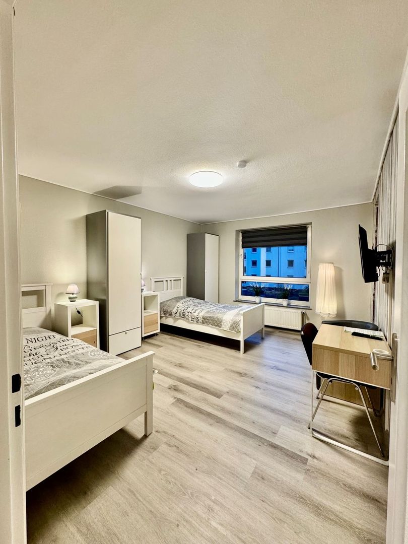 Schlicht eingerichtetes Schlafzimmer mit zwei Einzelbetten, Kleiderschrank, Schreibtisch mit Stuhl und Fenster mit Blick auf ein Nachbargebäude