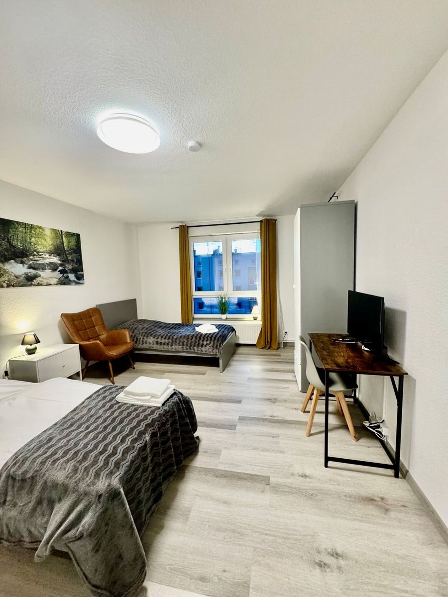 Schlicht eingerichtetes Zimmer mit zwei Betten, braunem Sessel, Schreibtisch mit Stuhl und Fernseher sowie Fenster mit gelben Vorhängen