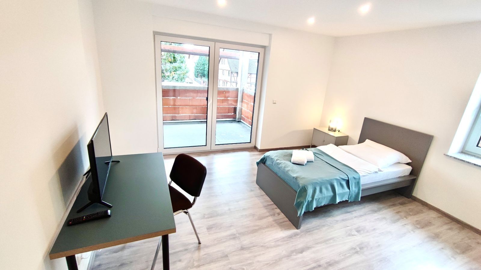 Helles Schlafzimmer mit Doppelbett, Schreibtisch mit Stuhl und großem Fenster mit Blick auf den Balkon